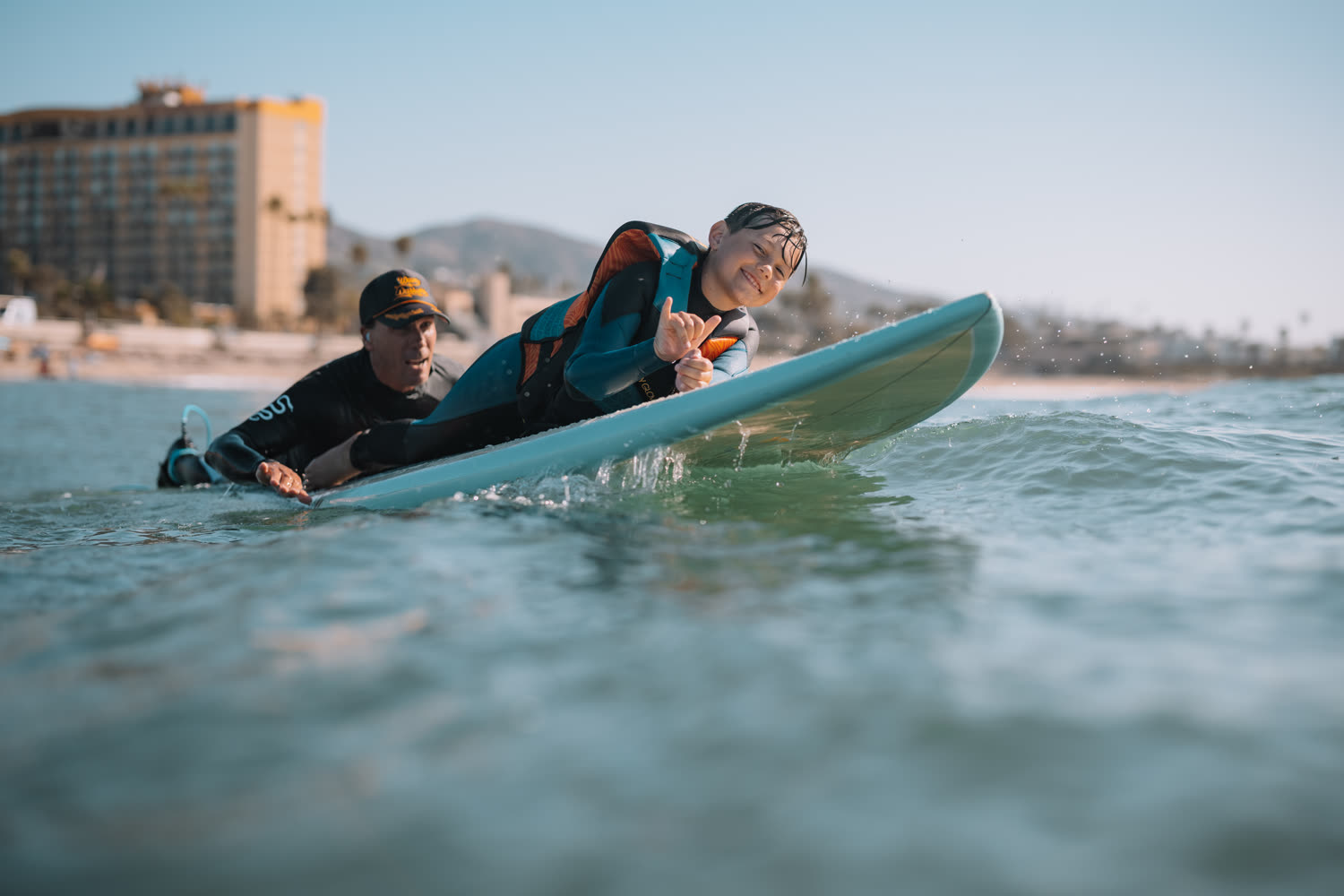 A young surfer throwing a shaka while lying on a board with an instructor alongside in the water