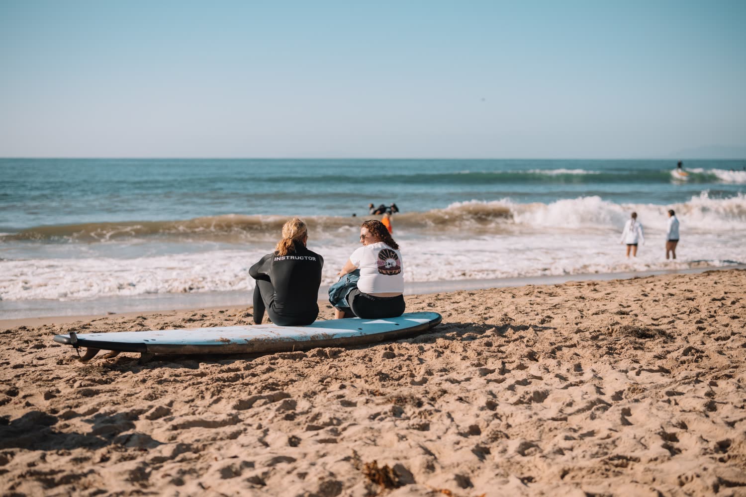 An instructor and a participant sitting on a surfboard on the sand, looking out at the waves