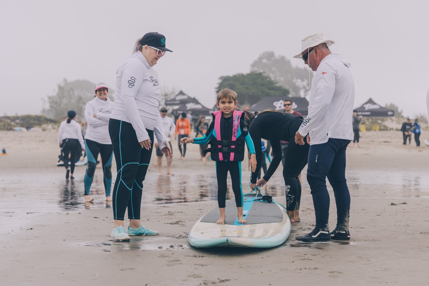 A young surfer stands on his own on a board on the sand, coaches beside him, ready for the water