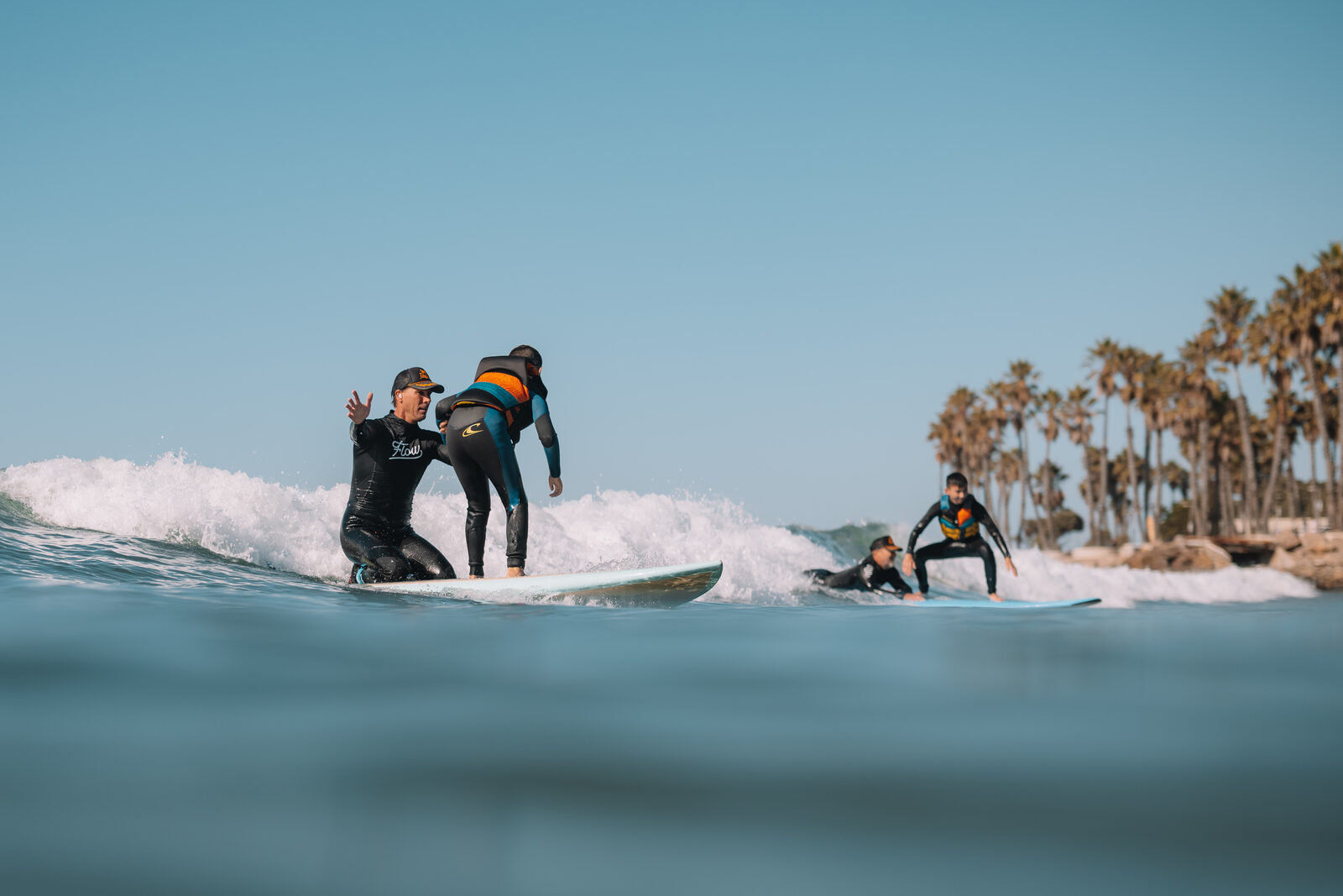 A Flow coach steadies a young athlete standing up on a wave while a second surfer rides behind them