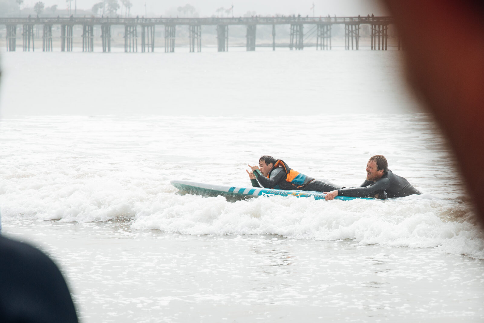 A young athlete throws a shaka on his first wave, lying on the board with his coach pushing from behind