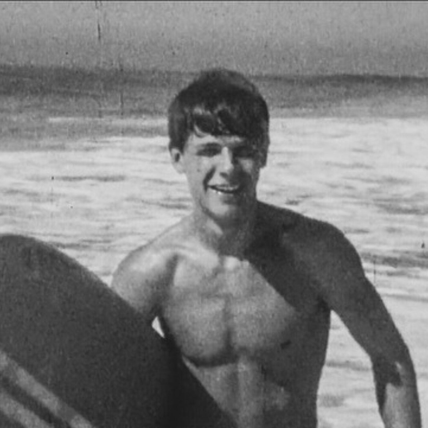 Pat's father as a young man, smiling with his surfboard at the water's edge in Malibu